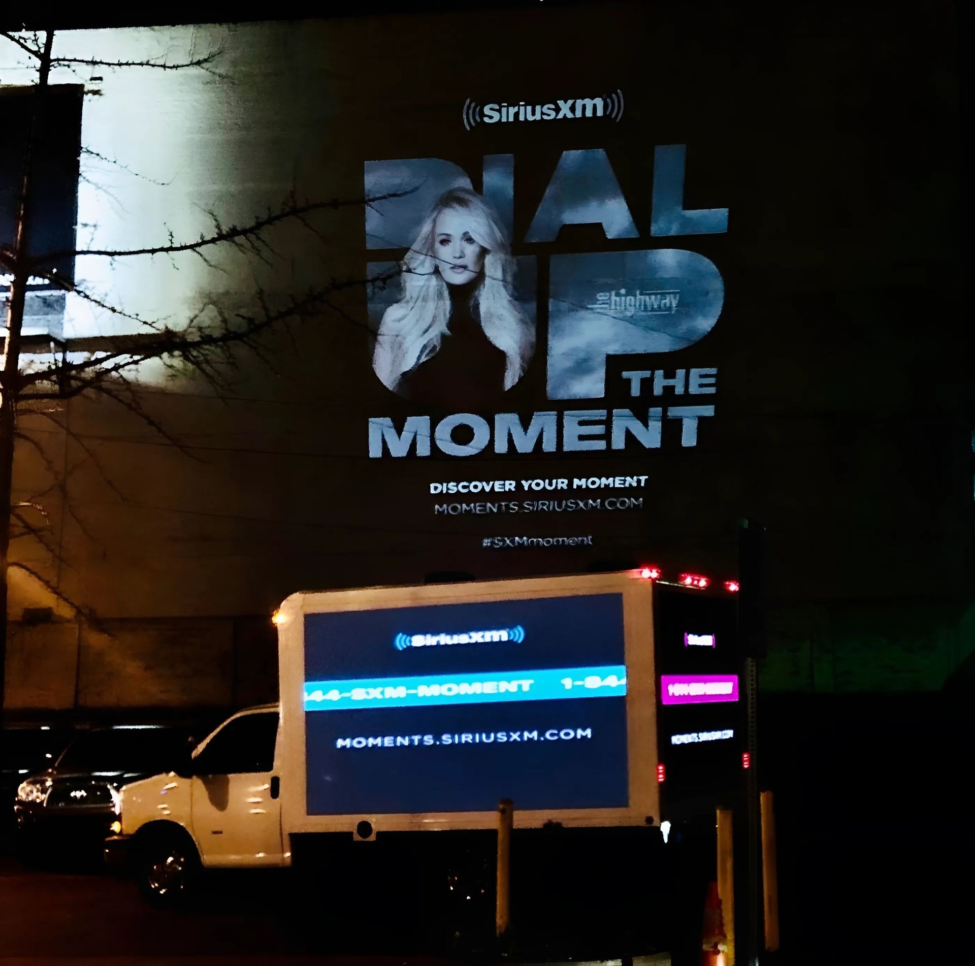 SiriusXM billboard at night with promotional message and contact information displayed on a city wall.